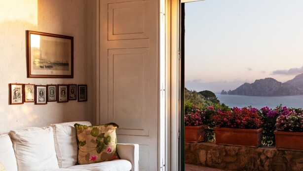 A room with a white sofa, a door leading to a balcony with a view of the sea and mountains, and framed pictures on the wall.