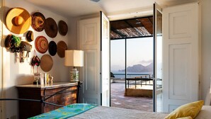 A bedroom with a bed, a desk, and a view of the sea through an open door.