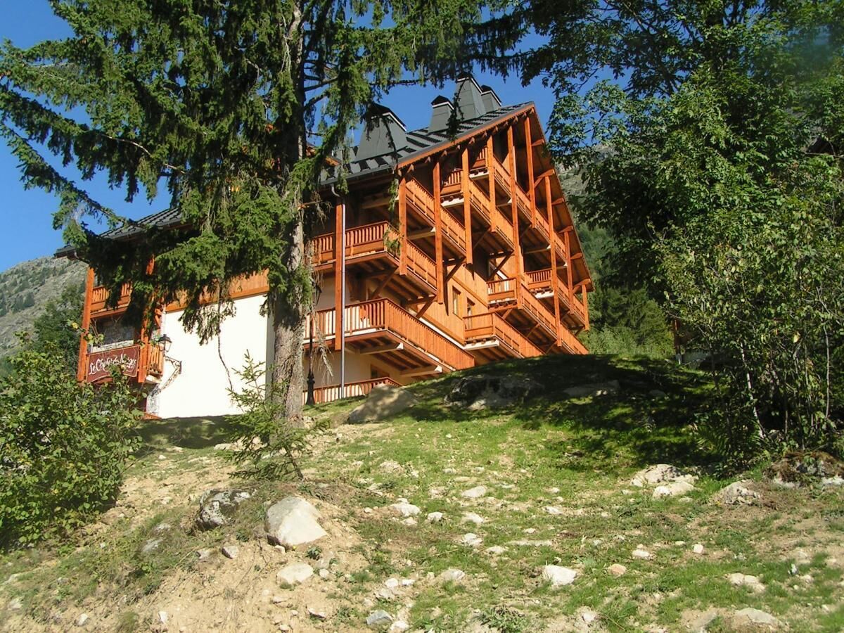 A wooden cabin with a balcony, surrounded by trees and a rocky hillside.