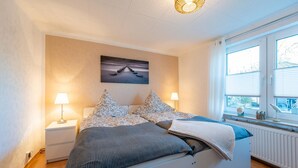 Ett sovrum med en säng, två sängbord, en lampa, en tavla på väggen och ett fönster med persienner.