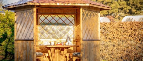 Un pabellón de madera con techo de tejas, una mesa con sillas y una ventana con celosía.