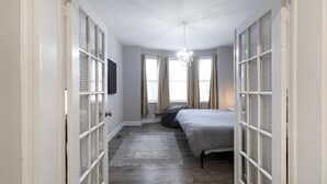 Un pasillo estrecho con una cama a la derecha, un ventanal con cortinas y un candelabro.