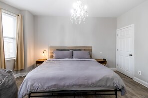 Ein Schlafzimmer mit einem Bett, Nachttischen, einer Chandelüster, einer Tür und einem Fenster mit Vorhängen.