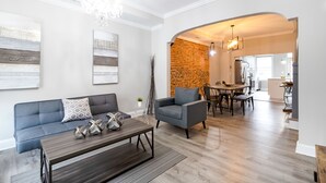 Una sala moderna con sofá, sillón y mesa de centro, un comedor con pared de ladrillo y una cocina al fondo.