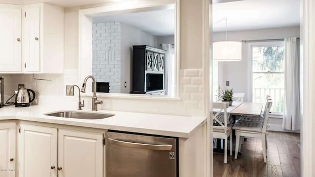 A kitchen with a sink, cabinets, and a dishwasher. There is a dining area with a table and chairs.