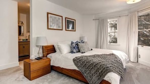 A bedroom with a wooden bed, white bedding, a grey blanket, two framed artworks, a bedside lamp, and a view of trees through a window.