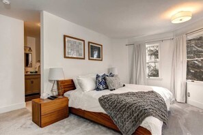 A bedroom with a wooden bed, white bedding, a grey blanket, two framed artworks, a bedside lamp, and a view of trees through a window.