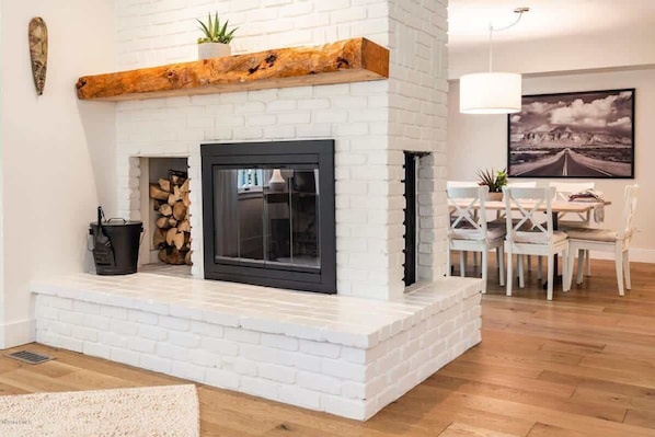 A modern living room with a white brick fireplace, a dining area with white chairs, and a large framed picture on the wall.