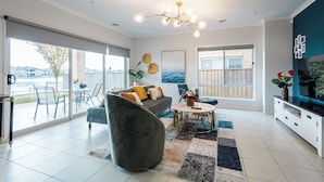 A modern living room with a grey sofa, a round coffee table, and a chandelier. There are large windows offering a view of the outdoors. - Luxury Waterfront Grand Villa in Melbourne (Point Cook)