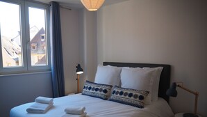 Un letto rifatto con lenzuola bianche e due asciugamani, una lampada da comodino e vista sugli edifici attraverso una finestra.