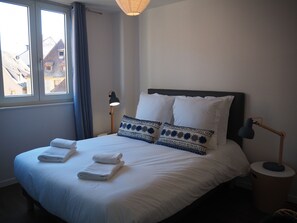 A neatly made bed with white linens and two towels, a bedside lamp, and a view of buildings through a window.