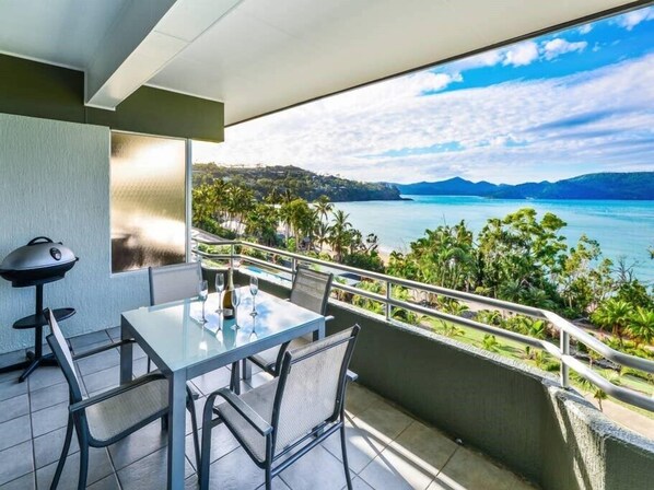 Un balcón con mesa y sillas, con vistas a la costa, montañas y palmeras.