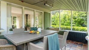 En overdækket terrasse med spisebord, fletstole og udsigt til frodig vegetation.
