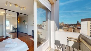 Una habitación de hotel moderna con balcón, piso de madera, cama con ropa blanca, zona de estar y vista a la ciudad.