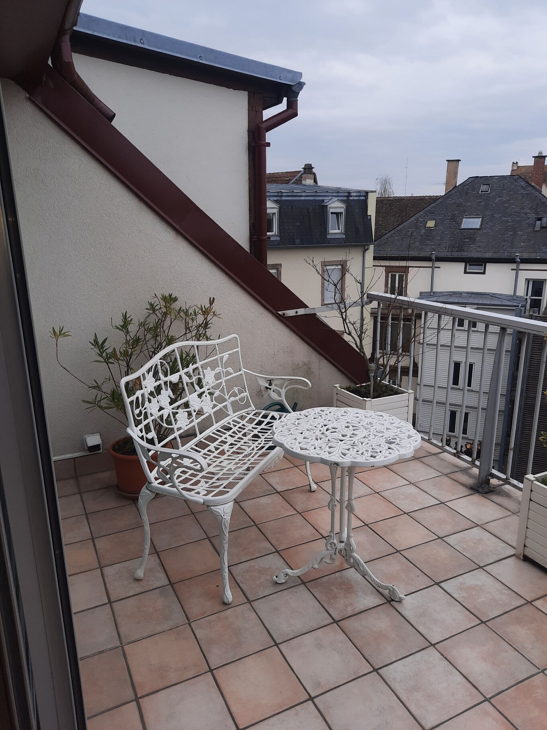 Un balcon avec un banc et une table en métal peints en blanc, des plantes en pot et une vue sur les bâtiments voisins.
