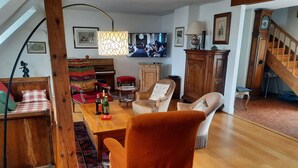 A cozy living room with a wooden dining table, a red armchair, and a sofa. A television is mounted on the wall, and there is a staircase leading to another level.