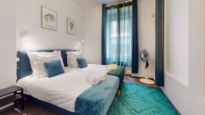 Un lit bien fait, avec une literie blanche et bleue, une table de chevet, un ventilateur sur pied et une fenêtre ornée de rideaux bleus.