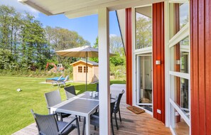 Relax on the covered deck with a table and chairs, surrounded by a well-manicured lawn and lush trees.