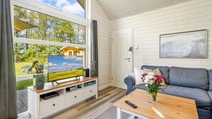 Ein Zimmer mit einem großen Fenster, einem Fernseher auf einem Holzbalken, einem grauen Sofa, einem kleinen Tisch mit Blumen und einer Tür mit einem gerahmten Bild an der Wand.