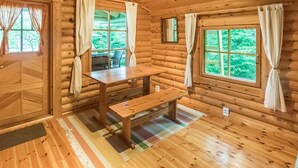 Uma cabana de madeira com interior rústico, uma mesa de jantar, bancos e janelas amplas com cortinas.