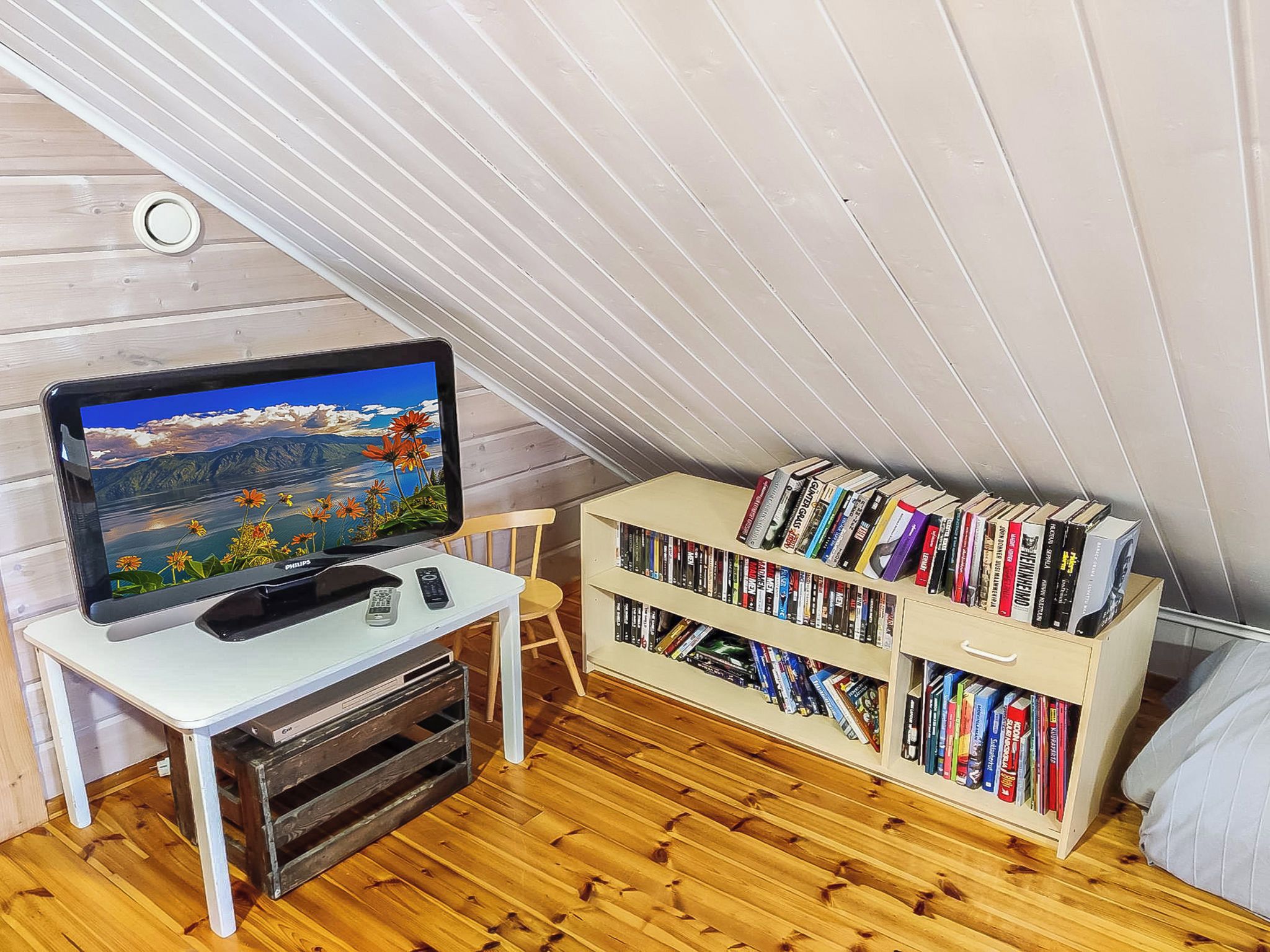 Een kamer met een televisie op een witte standaard, een boekenplank vol boeken en een houten vloer.