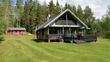 A two-story cabin with a balcony, surrounded by a well-maintained lawn and dense forest.