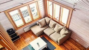 Um quarto aconchegante com piso de madeira, um sofá modular bege, uma mesinha branca e grandes janelas com molduras de madeira.