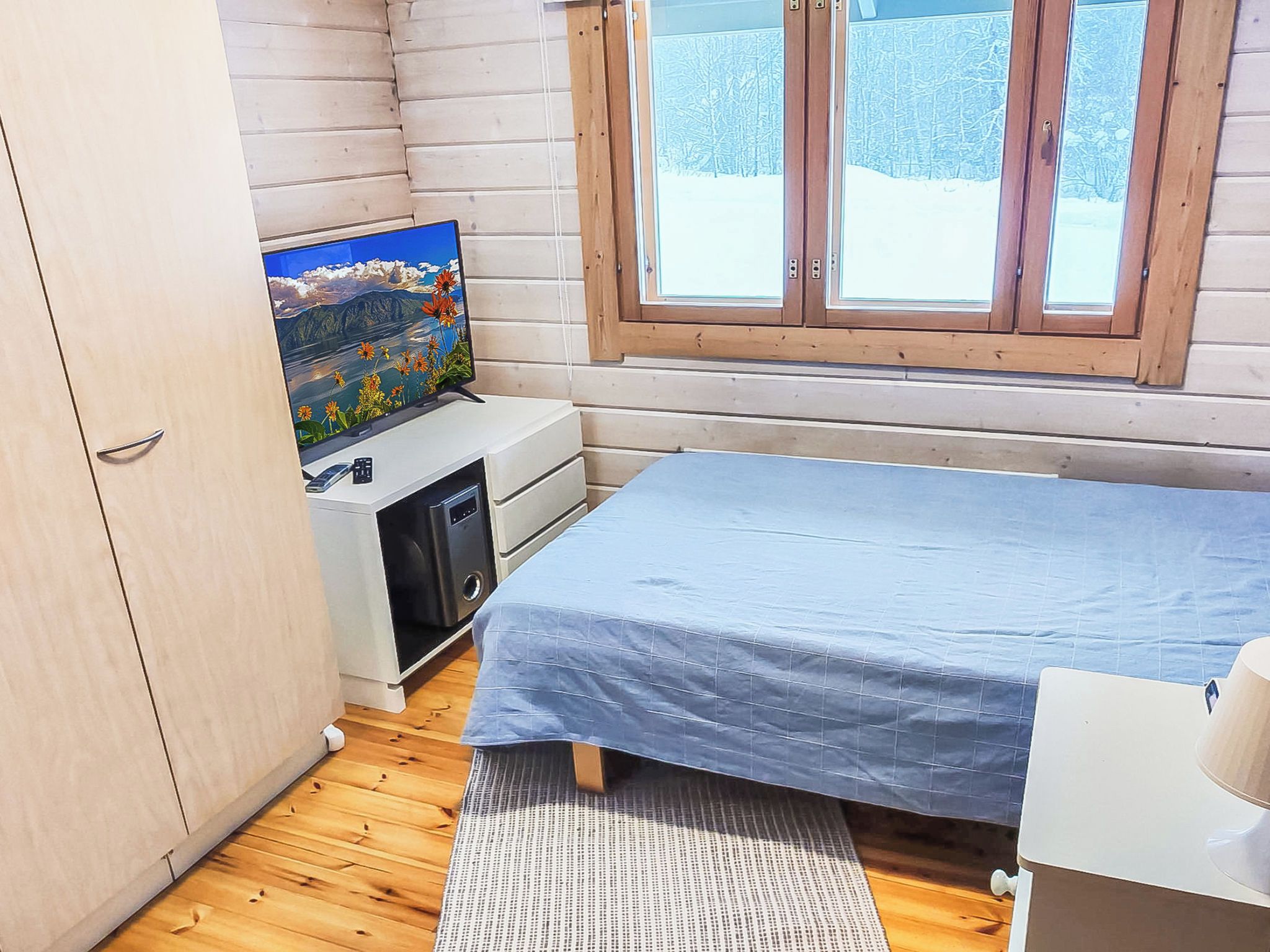 Een kleine kamer met een bed, een bureau met een computer, een televisie aan de muur en een raam met uitzicht op bomen.