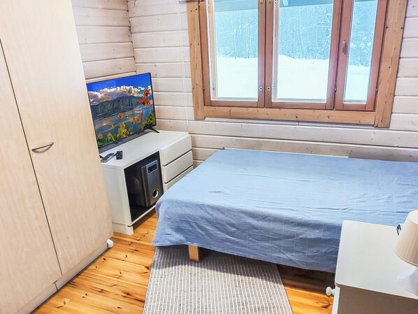 Une pièce de taille modeste comprenant un lit, un petit bureau avec un ordinateur, un téléviseur fixé au mur et une fenêtre donnant sur des arbres.