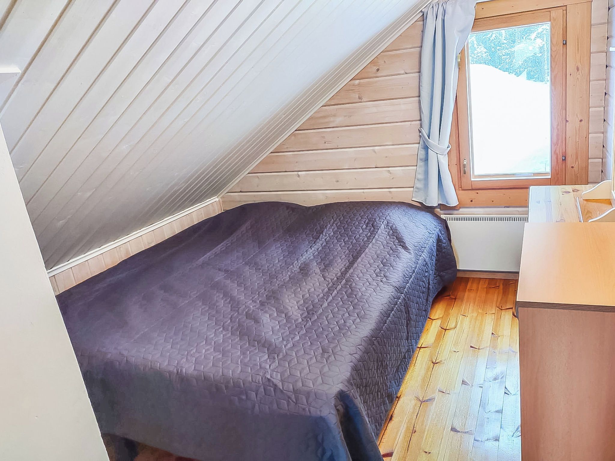 A small, single-bed room with a sloped ceiling, wooden walls, and a window with curtains.