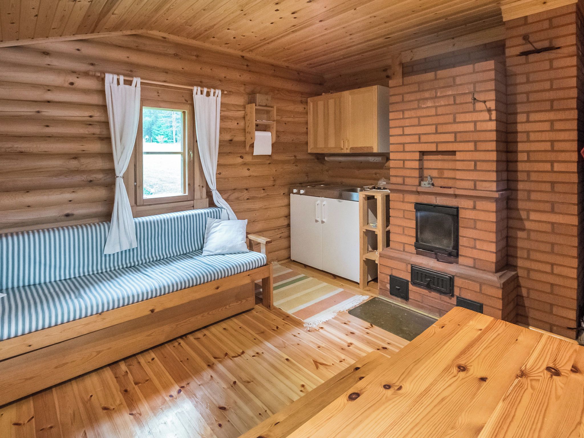 L’intérieur d’une cabane avec un plancher en bois, un canapé, un coin cuisine doté d’une cheminée en briques et une fenêtre avec des rideaux.
