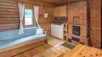 L’intérieur d’une cabane avec un plancher en bois, un canapé, un coin cuisine doté d’une cheminée en briques et une fenêtre avec des rideaux.