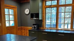 Une cuisine avec des meubles en bois, un four à micro-ondes, une horloge et de grandes fenêtres donnant sur la forêt.
