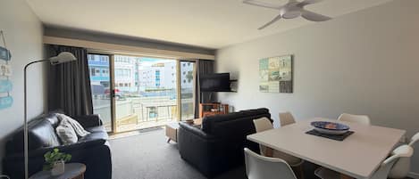 A modern living room with a sofa, a dining table with chairs, a ceiling fan, and a view through large windows.