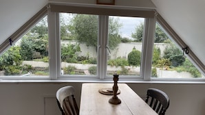 A dining area with a wooden table and chairs, a candle, and a view of a garden through large windows. - Pool Lodge, rural cottage-house, self catering property, rural, North Cornwall. (Helstone)