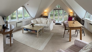 A cozy attic room with a loft bed, a wooden dining table, a sofa, and a coffee table with books. - Pool Lodge, rural cottage-house, self catering property, rural, North Cornwall. (Helstone)