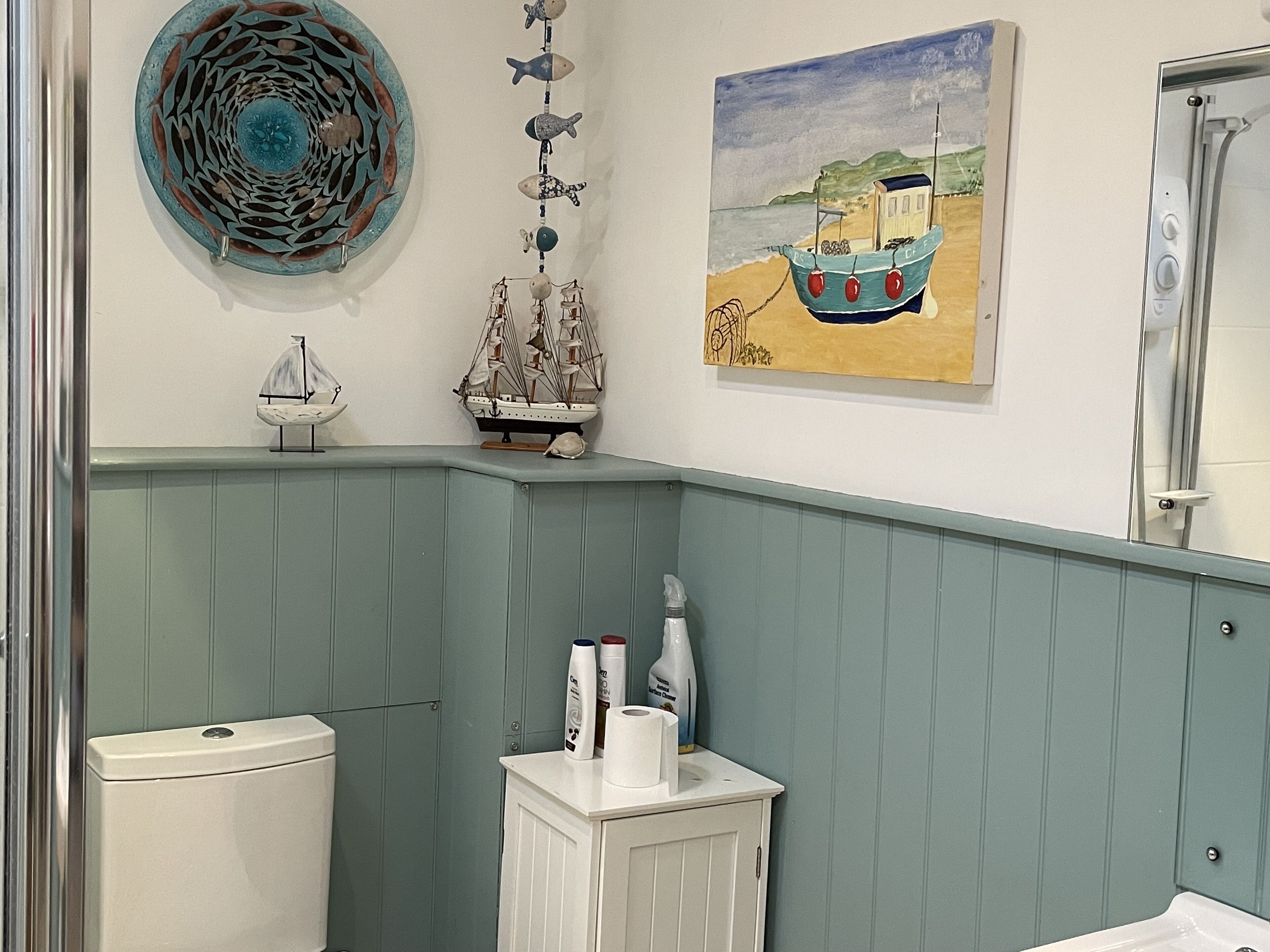 A bathroom with a toilet, a cabinet with toiletries, a decorative wall hanging, and a painting of a boat.