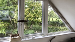 A room with a large window overlooking a garden, a small shelf with decorative items, and a ceiling fan. - Pool Lodge, rural cottage-house, self catering property, rural, North Cornwall. (Helstone)