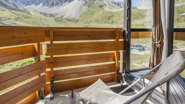 En balkon med gelænder i træ, en hvid stol og et bord med en kop. Der er bjergudsigt at se gennem den åbne dør.