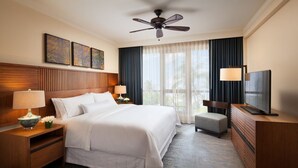 Ein Hotelzimmer mit einem großen Bett, zwei Nachttischen mit Lampen, einem Fernseher, einem Deckenventilator und einem Fenster mit Vorhängen.
