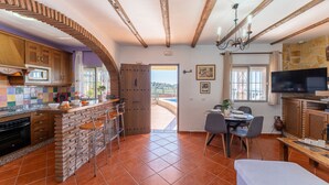 Una cocina con isla de piedra, taburetes de bar, zona de comedor con mesa y sillas, televisor y vistas a una terraza exterior por una puerta.