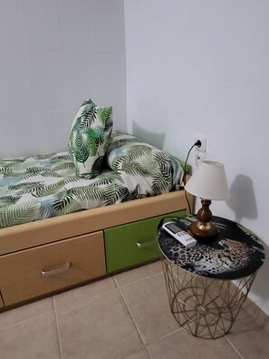 Ein kleiner Raum mit einem Bett, einer grünen Schublade, einem runden Tisch mit einer Lampe und einem Kissen mit Leopardenmuster.