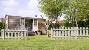 A wooden cabin with a porch, surrounded by a white picket fence, and a well-maintained lawn. - Secluded and Luxurious Shepherd's Hut (Melksham)