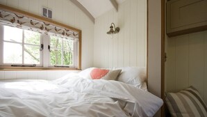 A bedroom with a bed, a window with curtains, a wall-mounted light fixture, and a door. - Secluded and Luxurious Shepherd's Hut (Melksham)