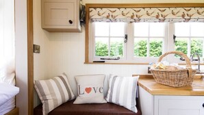 A cozy cabin interior with a wooden bench, a window with a view of greenery, and a kitchen area with a basket on the counter. - Secluded and Luxurious Shepherd's Hut (Melksham)