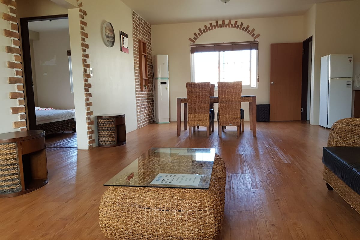 A spacious living area with a glass-top coffee table, wicker ottoman, and a dining table with chairs.