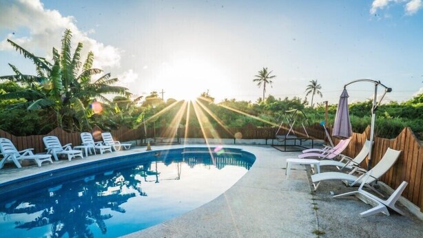 A swimming pool with lounge chairs and a swing set, surrounded by palm trees and a wooden fence. - Karis Villa (Saipan)