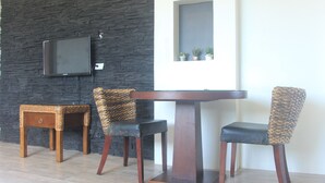 A dining area with a dark stone wall, a flat-screen TV mounted above a wooden table, and three chairs with woven backs. - Karis Villa (Saipan)