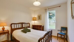 Een slaapkamer met een houten bed, een nachtkastje met een lamp, een stoel, een dressoir en een raam met uitzicht op groen.