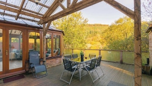 Eine überdachte Terrasse mit Tisch und Stühlen, ein Glastür-Eingang sowie Blick auf Bäume und Hügel.